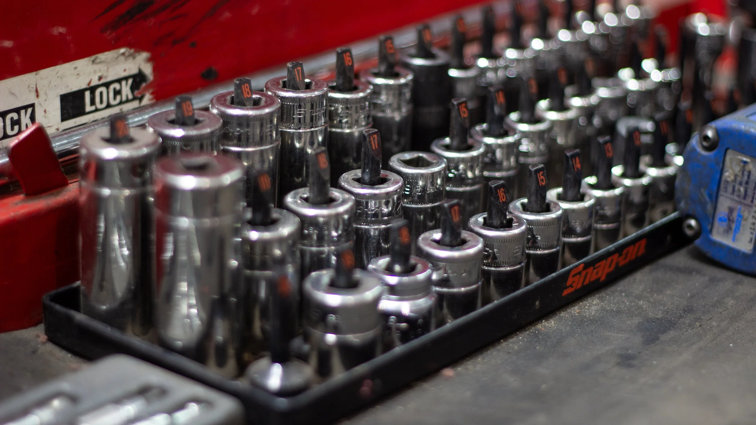 A set of metal sockets in a plastic toolbox drawer. Some sockets have numbered black and orange labels showing sizes like 15, 16, 17, 18, and 19. The toolbox is red with a label that says 'LOCK' in black and white.