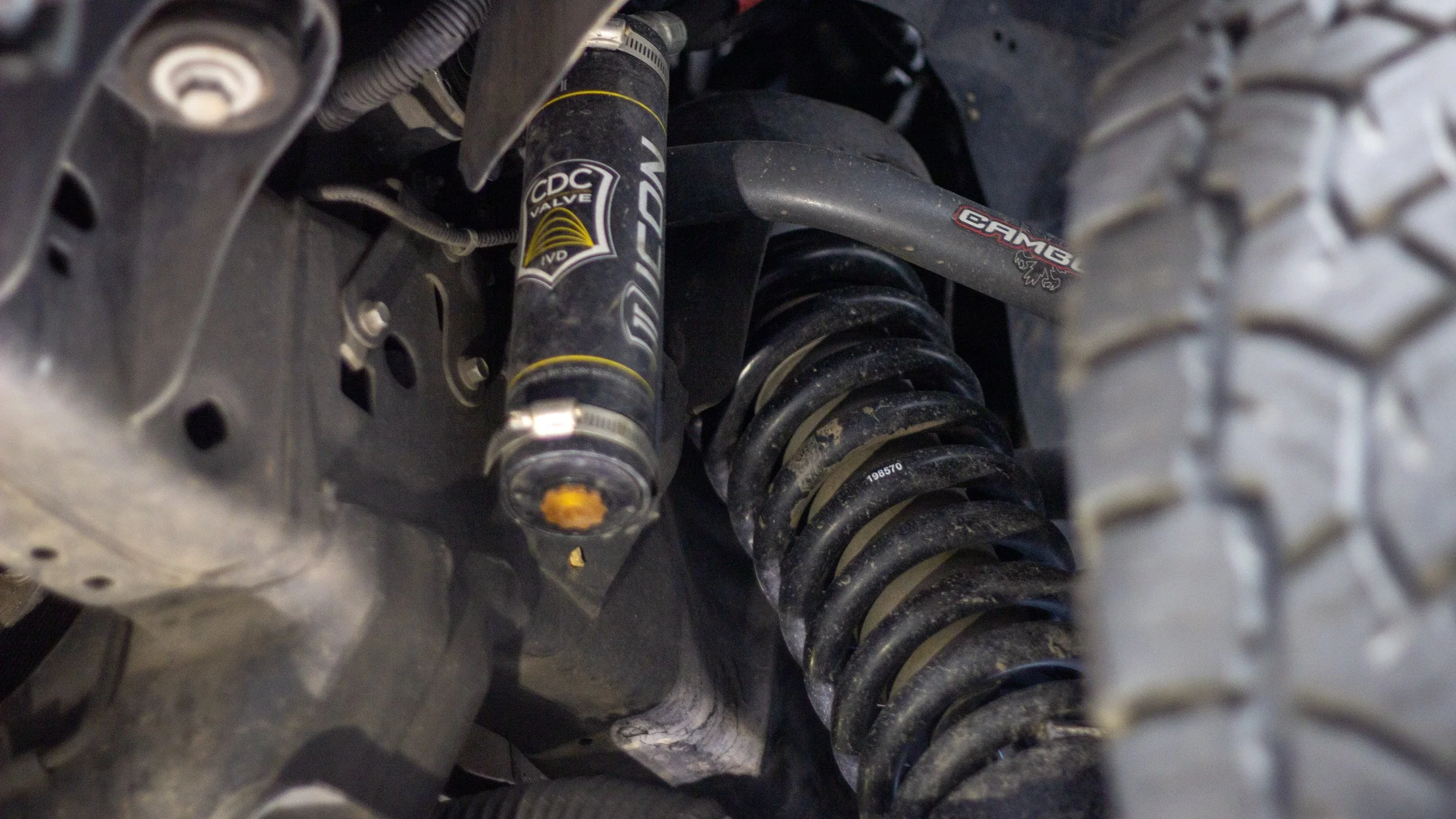 Close-up of a vehicle suspension system showing a shock absorber with the label CDC Valve, a coil spring, and part of the undercarriage and tire tread.