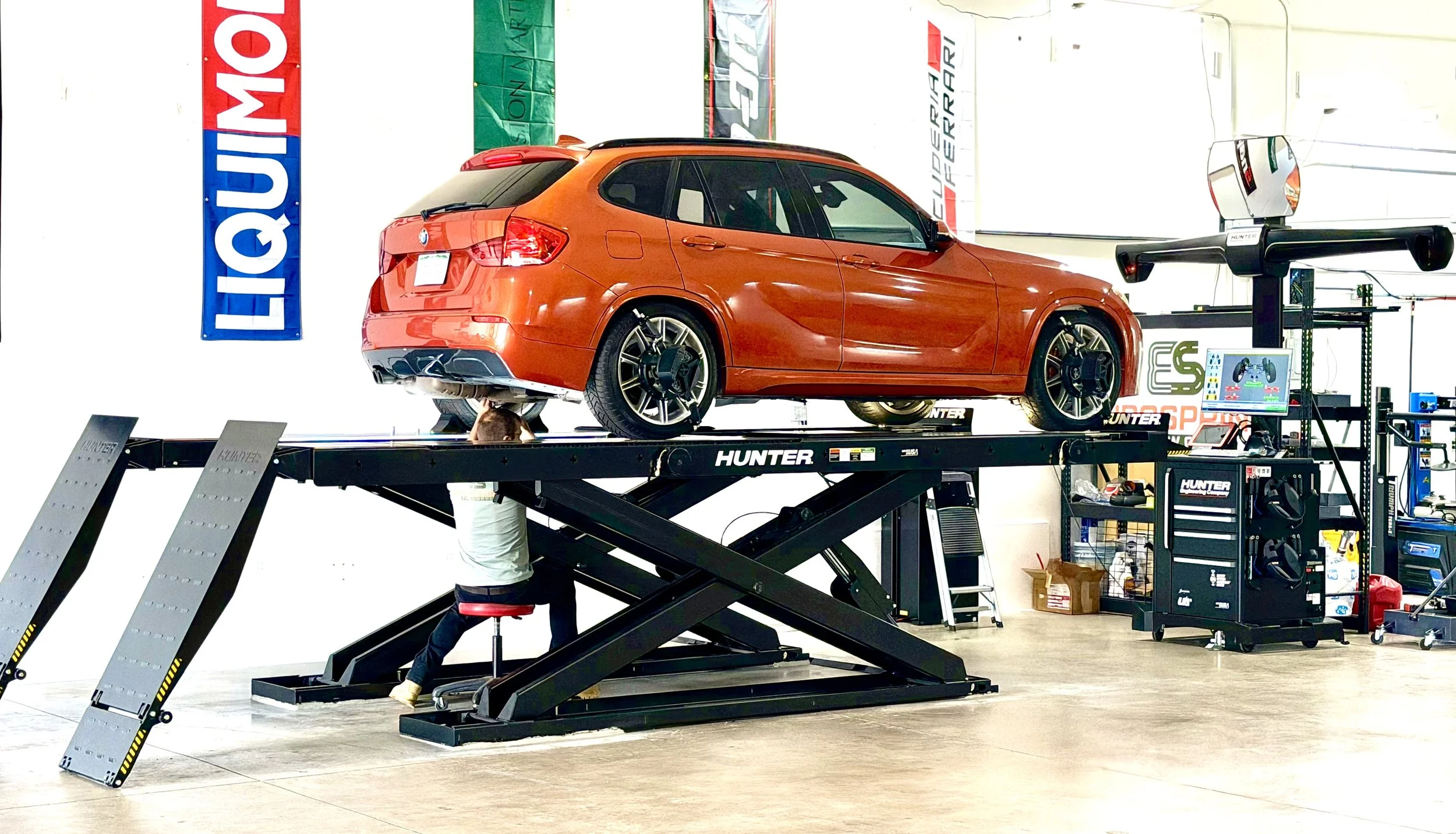 An orange BMW M model car is elevated on a car lift in a garage or workshop. The garage has banners with the logo 'LIQUI MOLY' and other automotive branding. Various tools and equipment are visible in the background.