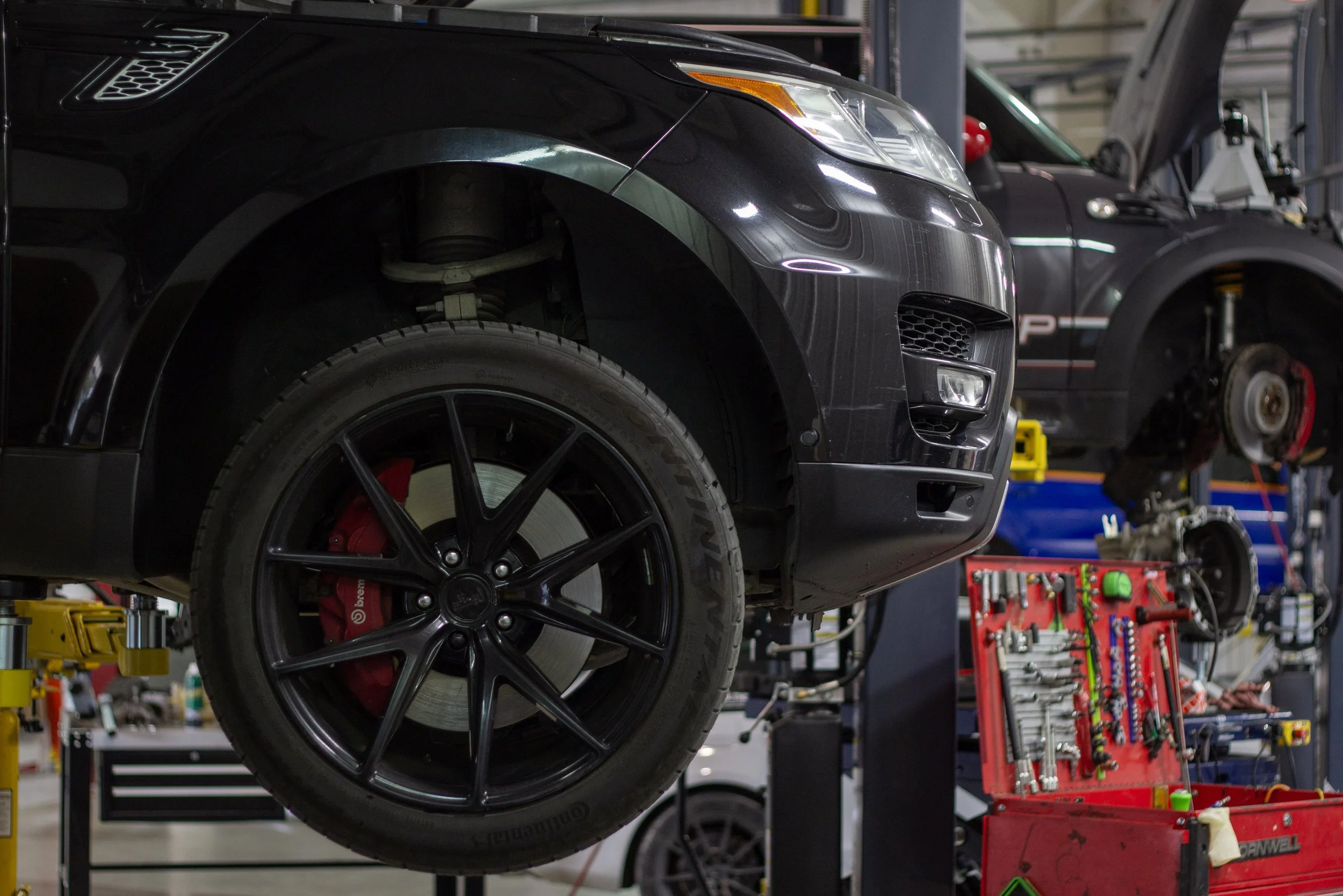 Close-up of a sleek black sports car on a lift in an auto repair shop, with a focus on the front wheel and tire.