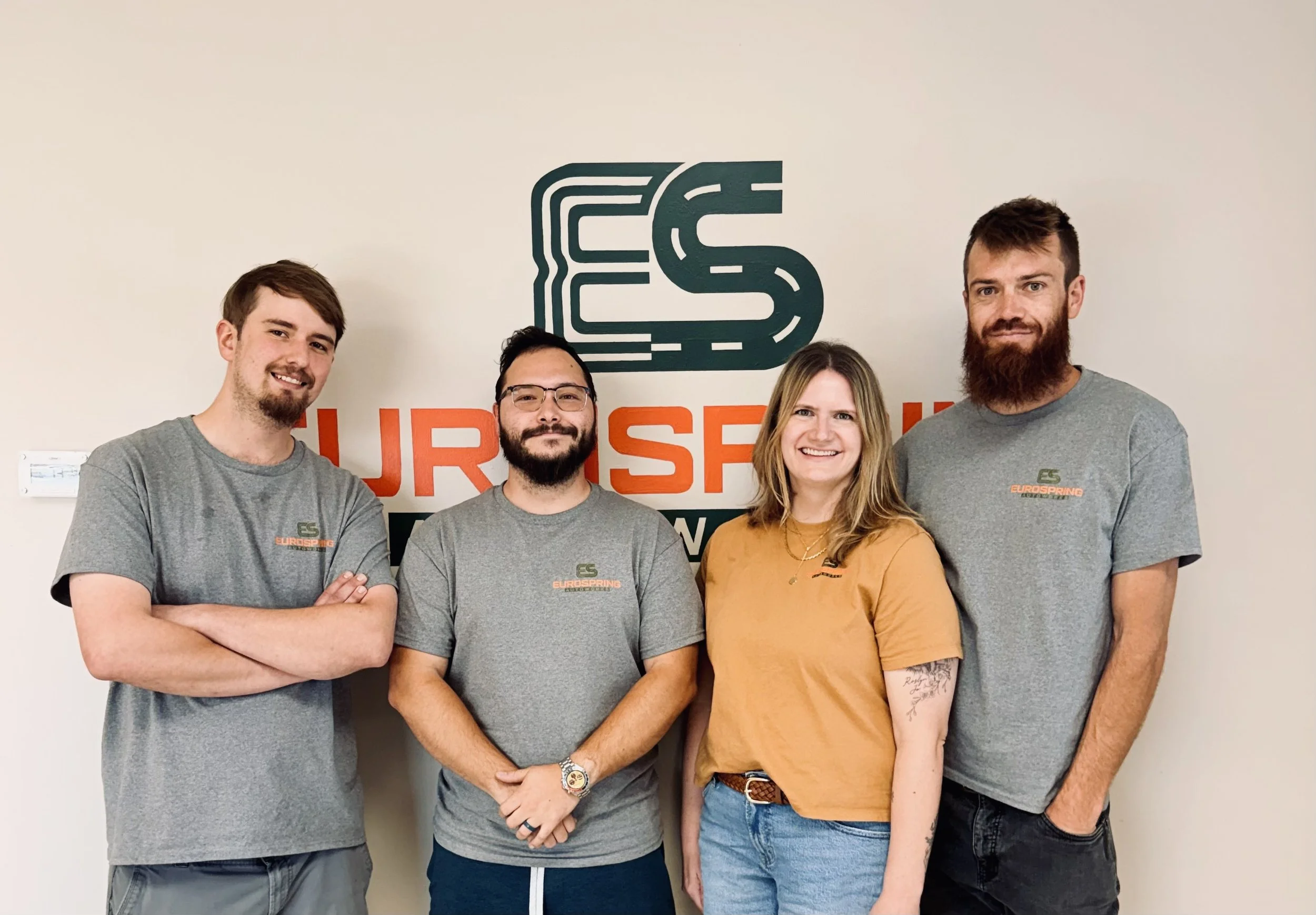 Four EuroSpring employees posing for a picture in front of the EUROSPRING painted logo wall. From the left it's Sam, Brandon, Laura, and Tyler.