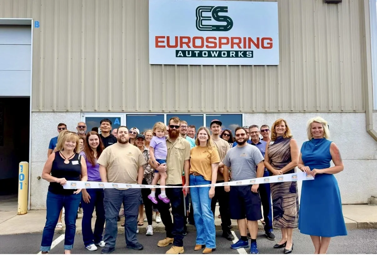 Group of people gathered in front of a building with a sign that reads 'EuroSpring Autoworks.' They are posing for a photo during a ribbon-cutting ceremony, with some holding a ribbon and others smiling.