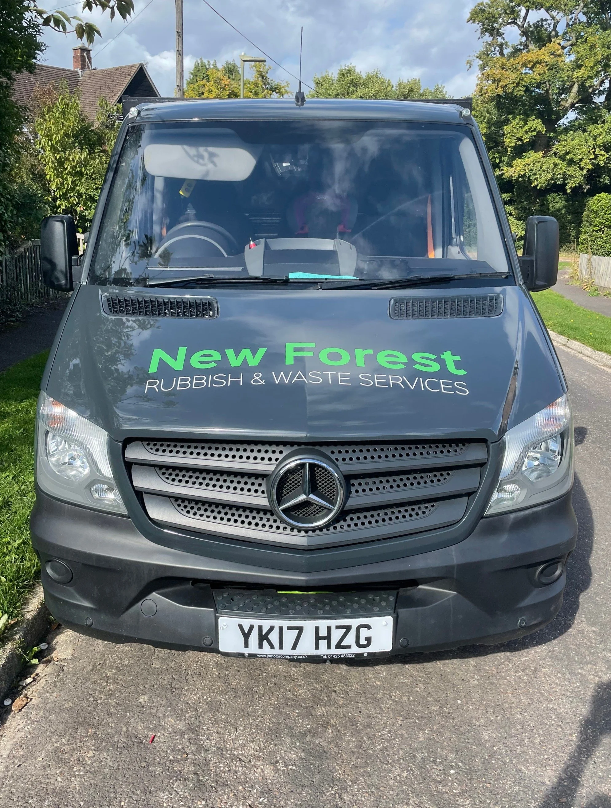Front view of a grey Mercedes-Benz utility vehicle with the sign 'New Forest Rubbish & Waste Services' on the hood, parked on a paved surface with trees and buildings in the background.