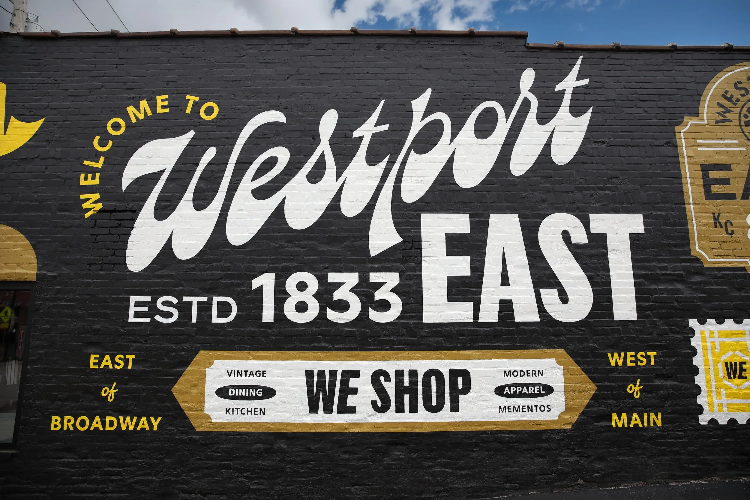 A mural on a brick wall welcoming visitors to Westport East, established in 1833, with details about the East and West side of the area, featuring a vintage and modern style, and mentioning a shop that sells food, apparel, mementos, and more.