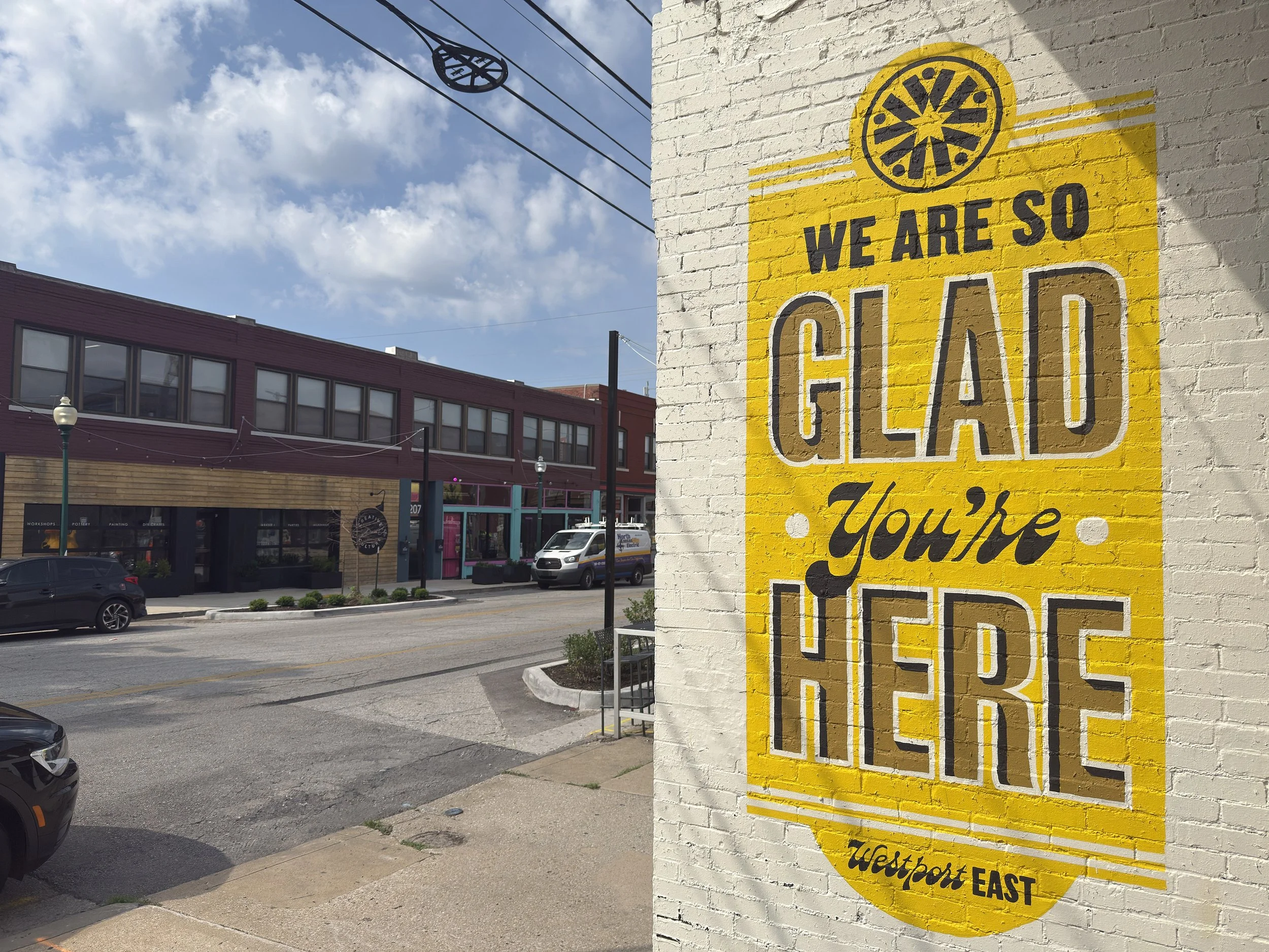 A yellow sign on a white brick wall that reads 'WE ARE SO GLAD You're HERE' with a decorative circular logo at the top. Westport East