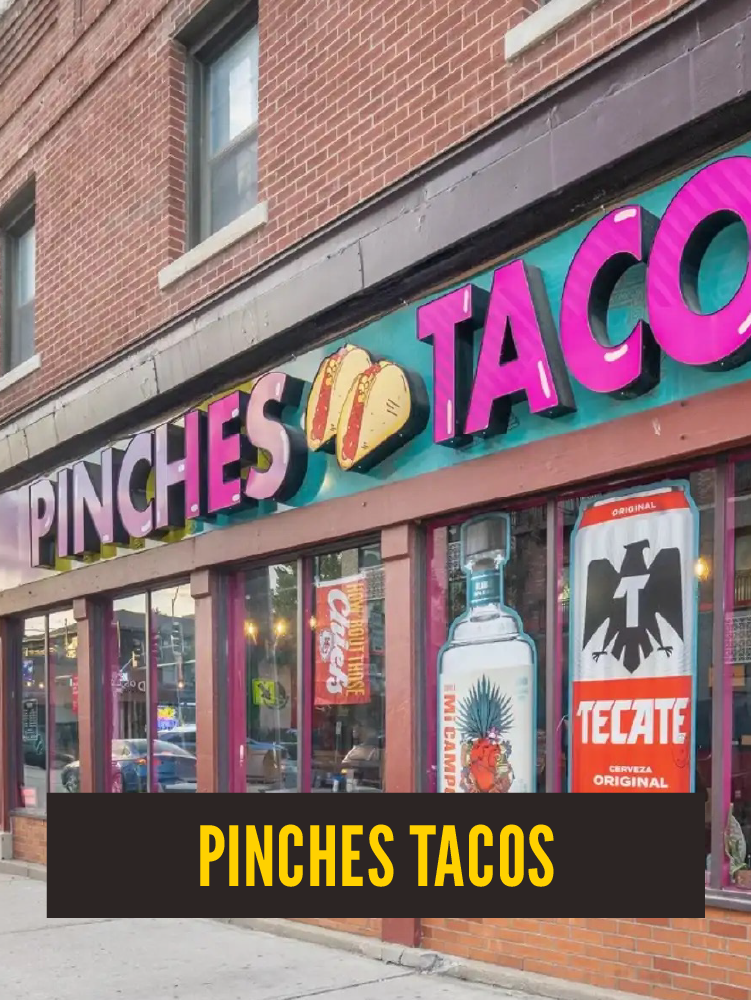 The storefront of Pinches Tacos, a taco restaurant, with the store name in large pink letters and a taco graphic between the words. The window displays a bottle of tequila and a Tecate beer advertisement.  Westport East.