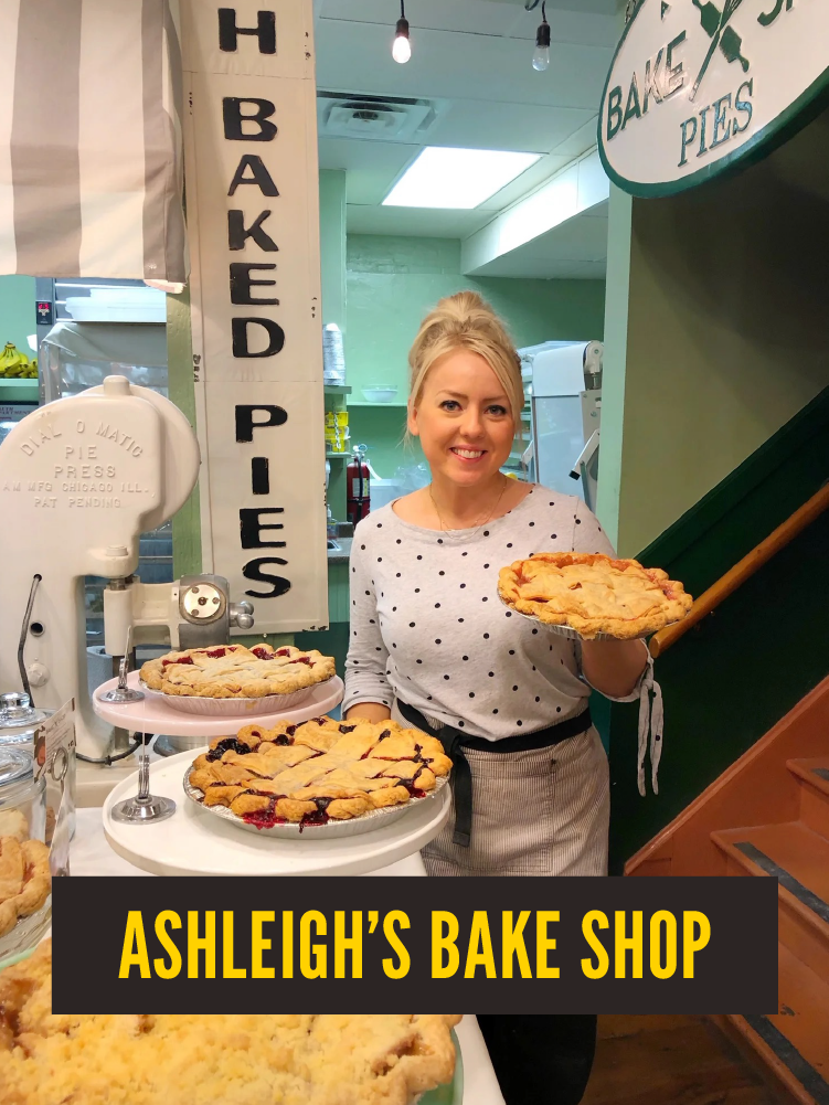 A woman standing behind a display of pies at Ashleigh's Bake Shop, holding a pie in her hand and smiling. The shop's sign is visible, along with various baked pies on stands. Westport East.