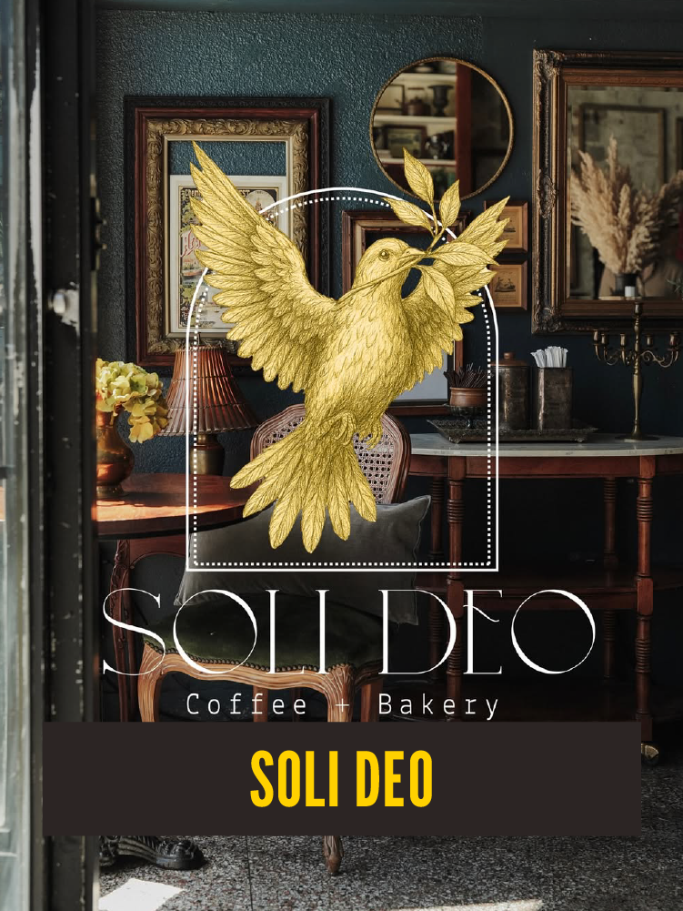Interior of a cozy coffee and bakery shop called Soli Deo, featuring a decorative golden dove with leaves, framed artwork on dark teal walls, a wooden table with a lamp and flowers, and a mirror reflecting the room.  Westport East.