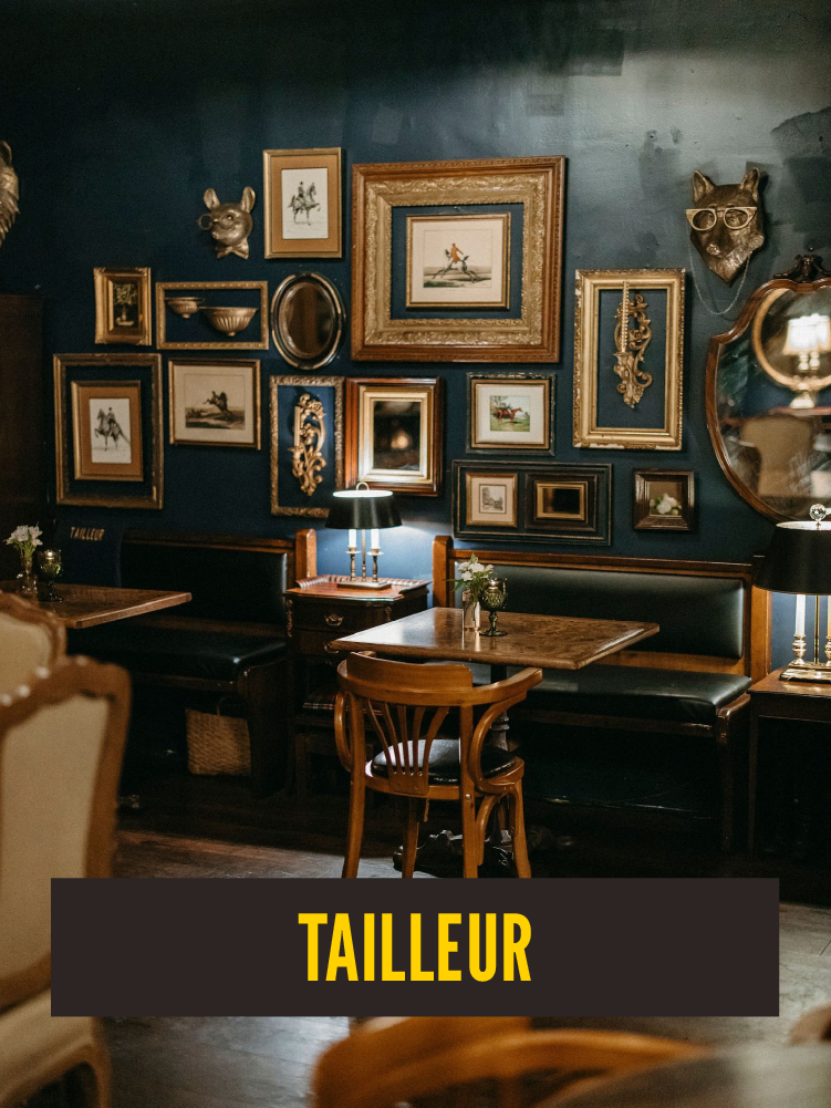 Interior of a dimly lit room with a gallery wall featuring framed artwork and decorative items. There are tables with small flower arrangements, lamps, and vintage furniture.  Westport East. Taileur.