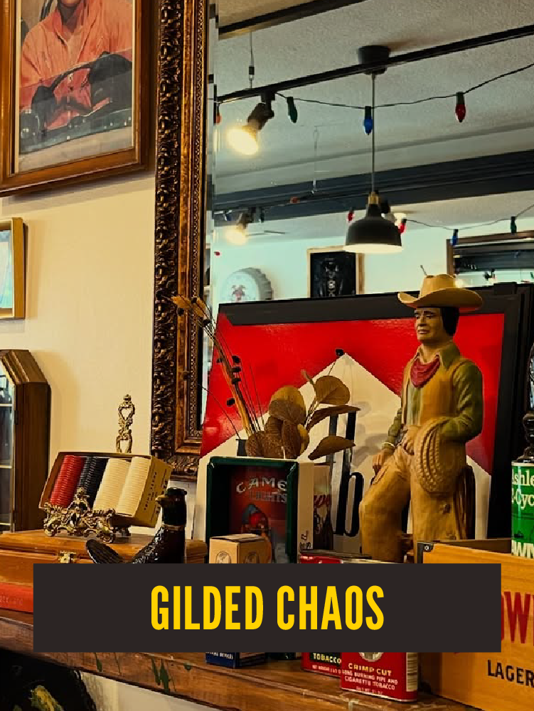 Decorative scene featuring a cowboy figure, a mirror, and various small collectibles on a wooden surface, with framed artwork and hanging lights in the background.  Westport East. Gilded Chaos.