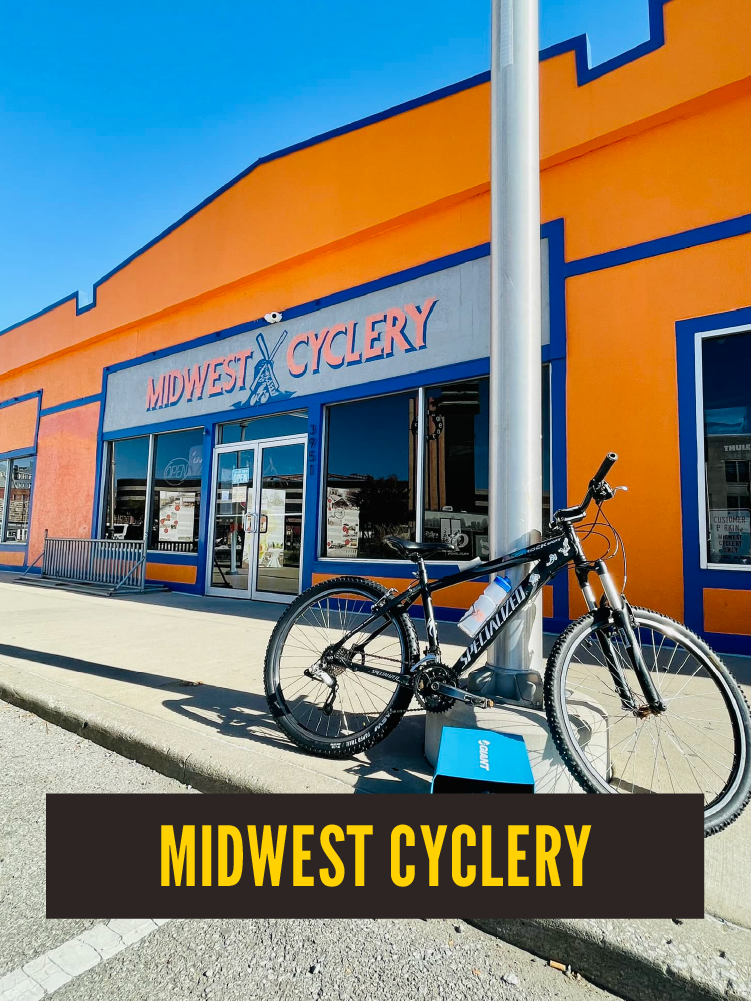 A storefront with an orange and blue exterior that reads 'Midwest Cyclery.' A mountain bike is locked to a pole in front of the store, with a blue donation box or display at its base. The sky is clear and blue.  Westport East. Main Street.
