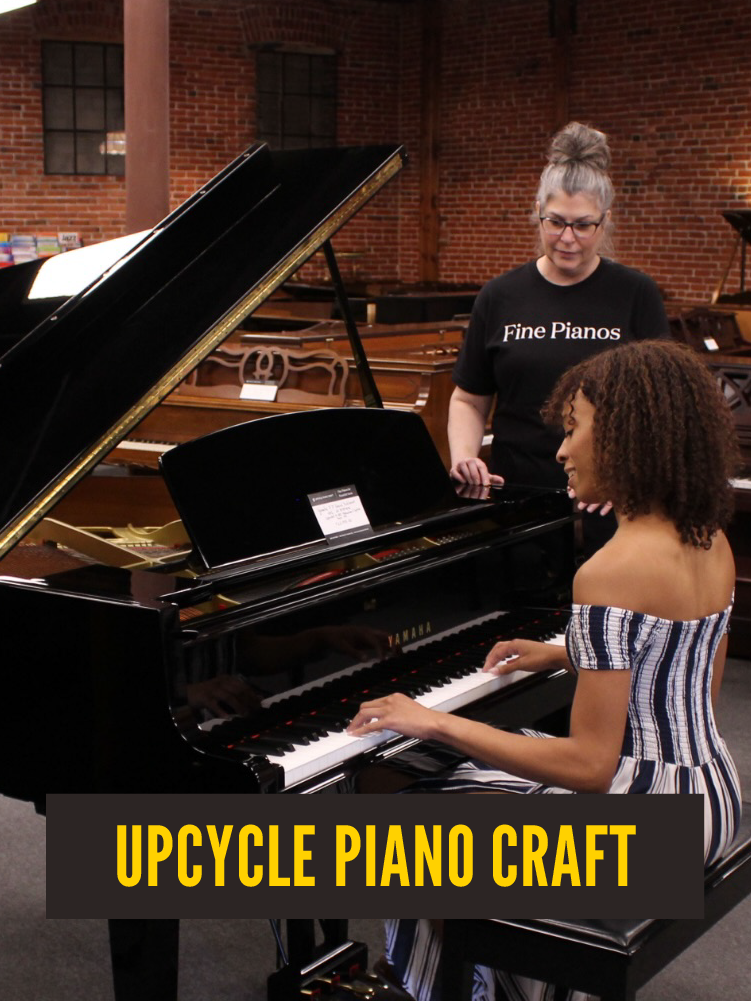 Yamaha upright piano, while another woman with glasses and gray hair looks on, in a piano store with brick walls and other pianos in the background. 'Fine Pianos'. Upcycle Piano Craft.  Westport East. Main Street.