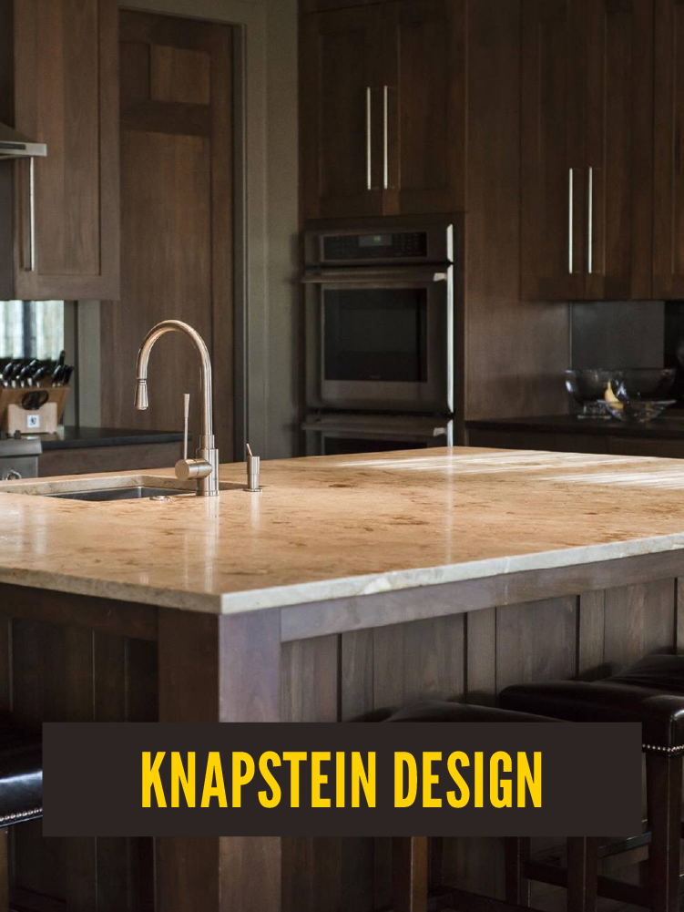 A modern kitchen with dark wood cabinets, a marble island countertop, a stainless steel sink, and a double oven in the background. There are black stools at the island and bowls on the counter. Knapstein Interior Design.  Westport East.