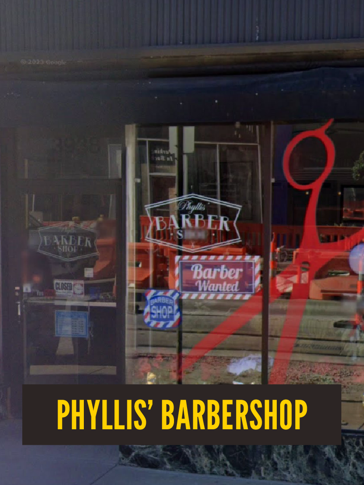 The storefront of Phyllis' Barbershop features signs that say 'Barber Shop,' 'Barber Wanted,' and 'Closed,' along with a large red barber pole logo. Inside, there's a glimpse of the interior with chairs and shelves.  Westport East. Main Street.