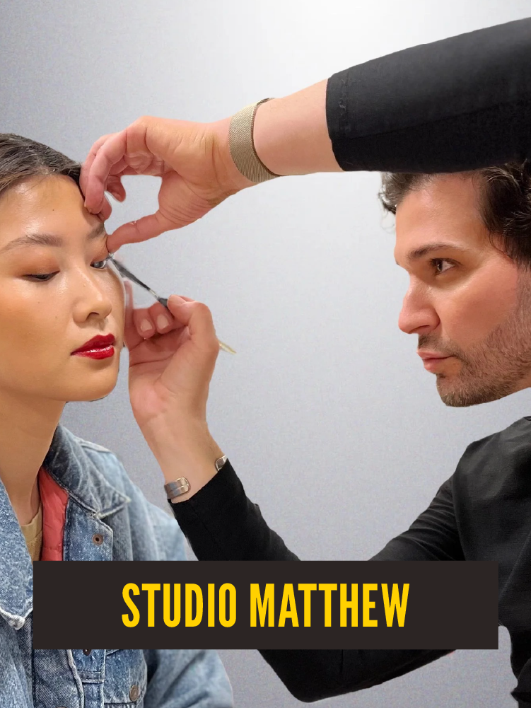 Makeup artist applying makeup to young woman with bold red lipstick, wearing denim jacket, with text overlay 'STUDIO MATTHEW'.  Westport East.
