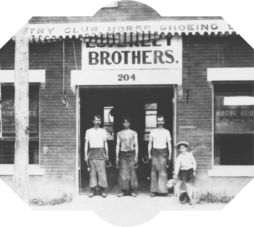 Black and white photo of three men, a boy, and a dog standing in front of a brick building with signs that read "Luxury Brothers" and "Horse Shoeing". Old Westport. Esterley Blacksmith Shop, 1906