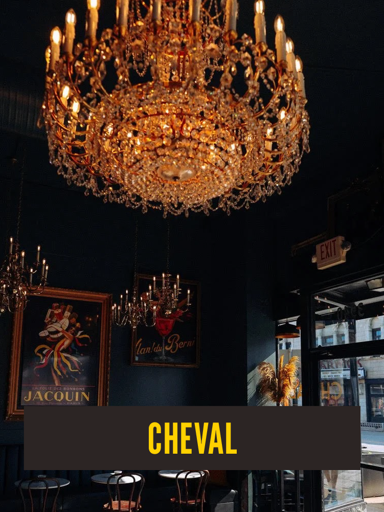 Interior of a dimly lit room with a large ornate chandelier hanging from the ceiling, framed artwork on the wall, small tables and chairs, and an exit sign near the door, with the word "CHEVAL" displayed at the bottom.  Westport East.