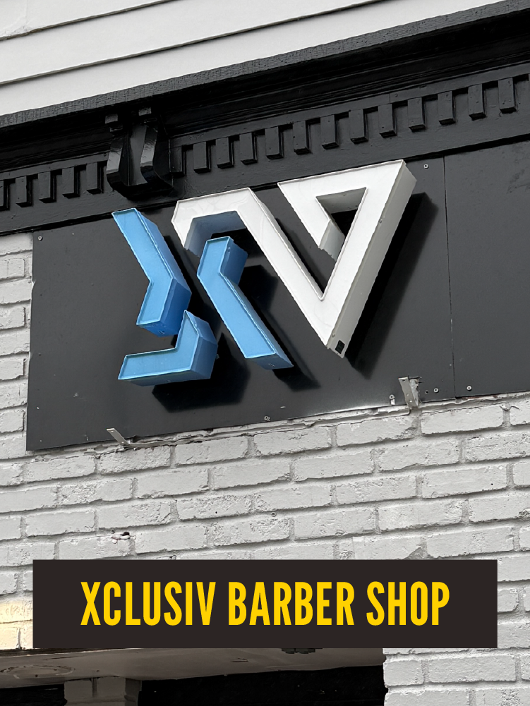 Sign for Xclusive Barber Shop with stylized 3D letters in blue and white, mounted on a brick wall.  Westport East.