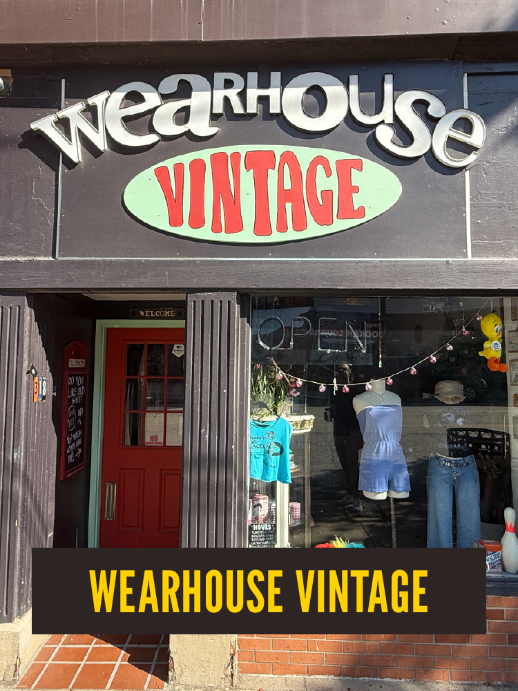 Storefront for Wearhouse Vintage featuring a large sign with the store name and a green oval with red text reading 'VINTAGE.' The display window shows mannequins, clothing, and decorations.  Westport East.