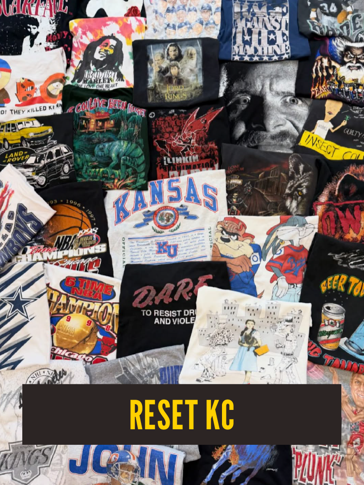 A collection of various colorful t-shirts with pop culture, sports, music, and artwork designs, including references to Kansas City, with a black banner at the bottom that reads "RESET KC" in yellow letters.  Westport East.
