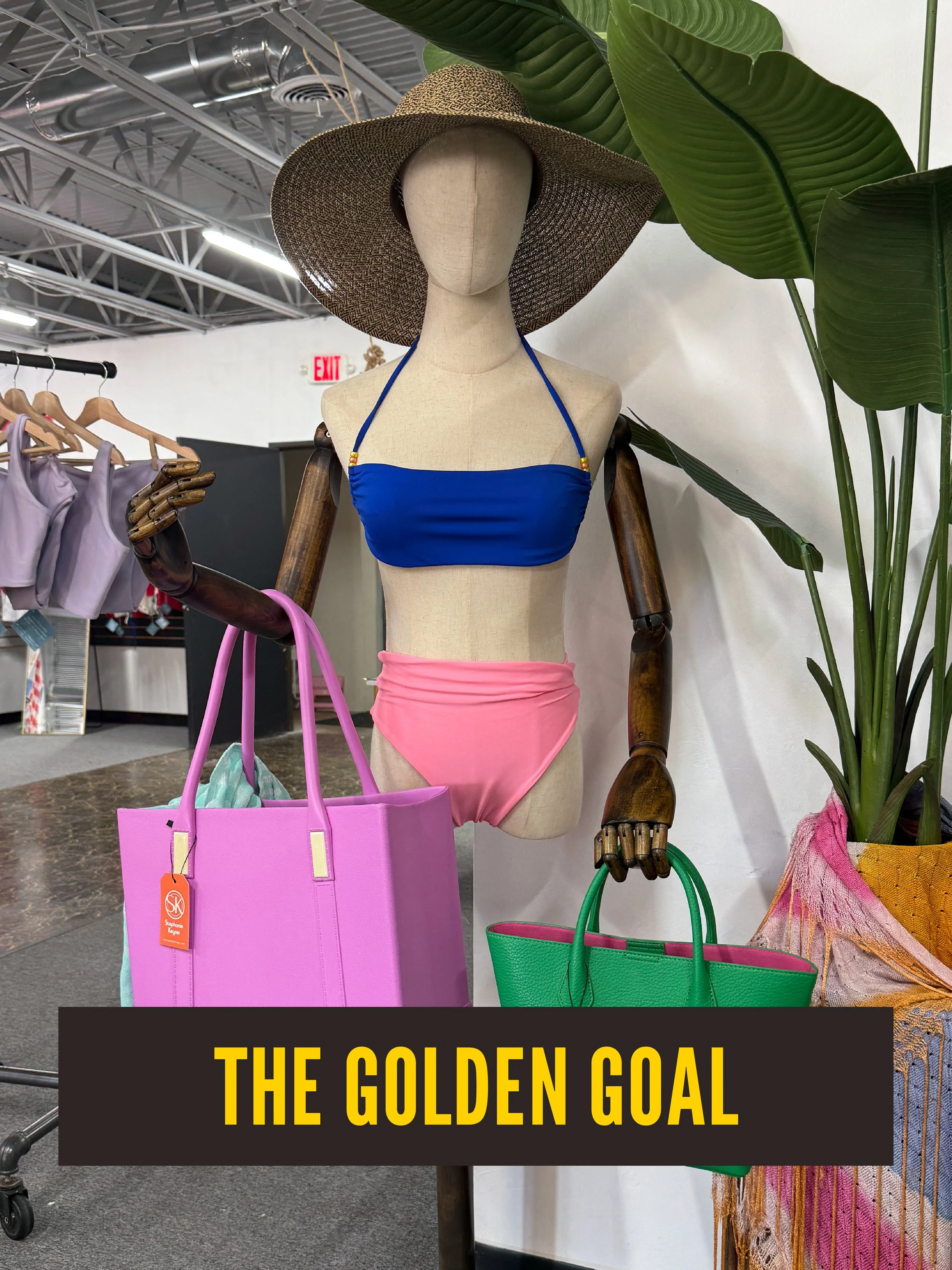 Display of a mannequin wearing a large woven sun hat, a custom swimsuit, surrounded by colorful tote bags, with large green tropical leaves in the background and a sign reading "The Golden Goal".  Westport East.
