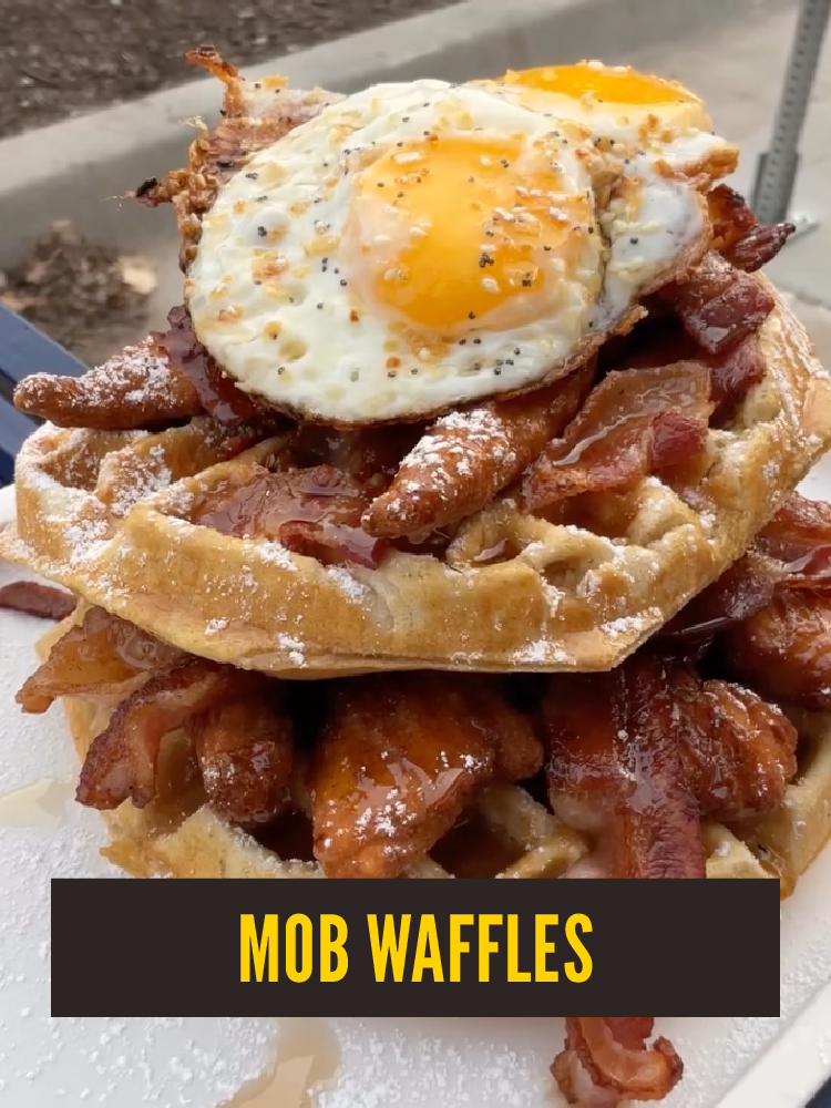 A stack of waffle-shaped fried chicken topped with bacon and two sunny-side-up eggs, garnished with syrup and sprinkled with powdered sugar.  Westport East.