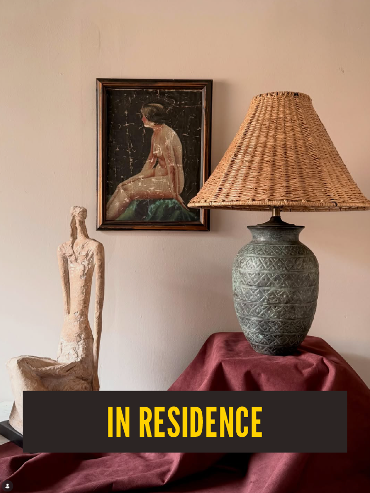 Decorative room with a framed painting of a woman sitting, a large patterned table lamp with a wicker shade, a carved statue of a seated figure, and a table draped with a red cloth. The text "In Residence" appears at the bottom. Westport East.