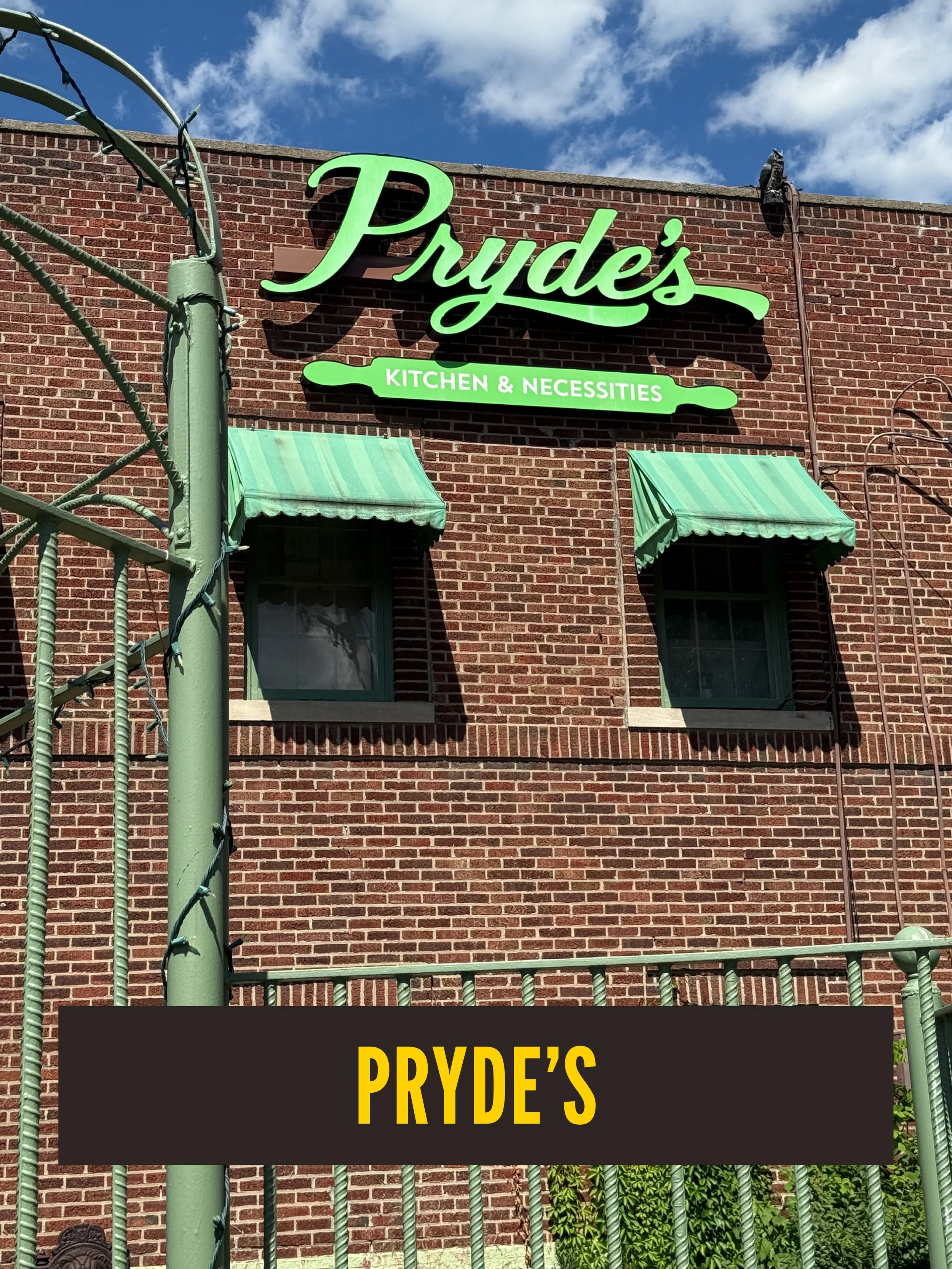 The image shows a brick building with green awnings above two windows. A neon green sign reads 'Pryde's Kitchen & Necessities'. A green metal fence and a utility pole with wires are in the foreground. Wesport East. Wesport Road.