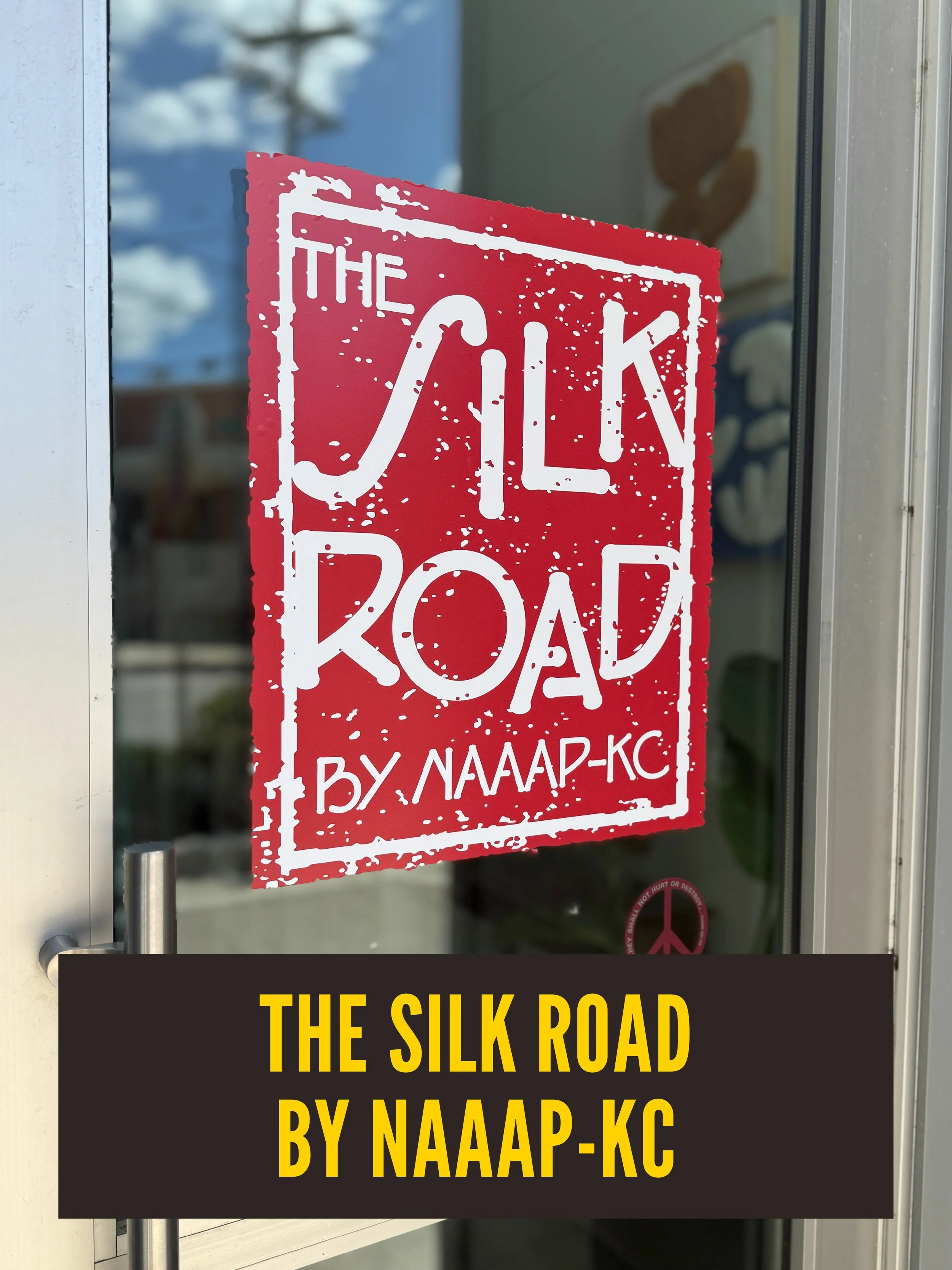 A red and white sign on a glass door reads 'THE SILK ROAD BY NAAA-KC' in bold letters. The background shows a reflection of a cloudy sky and some blurred objects inside the building.  Westport East.