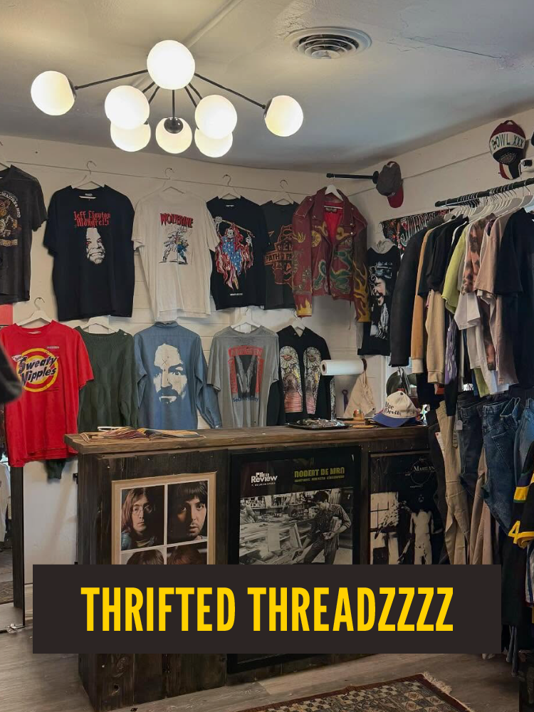 Inside a thrift store with various band and pop culture t-shirts hanging on the wall, a wooden counter with framed pictures and posters, and a ceiling light fixture with multiple bulbs.  Westport East. Thifted Threadzzzz