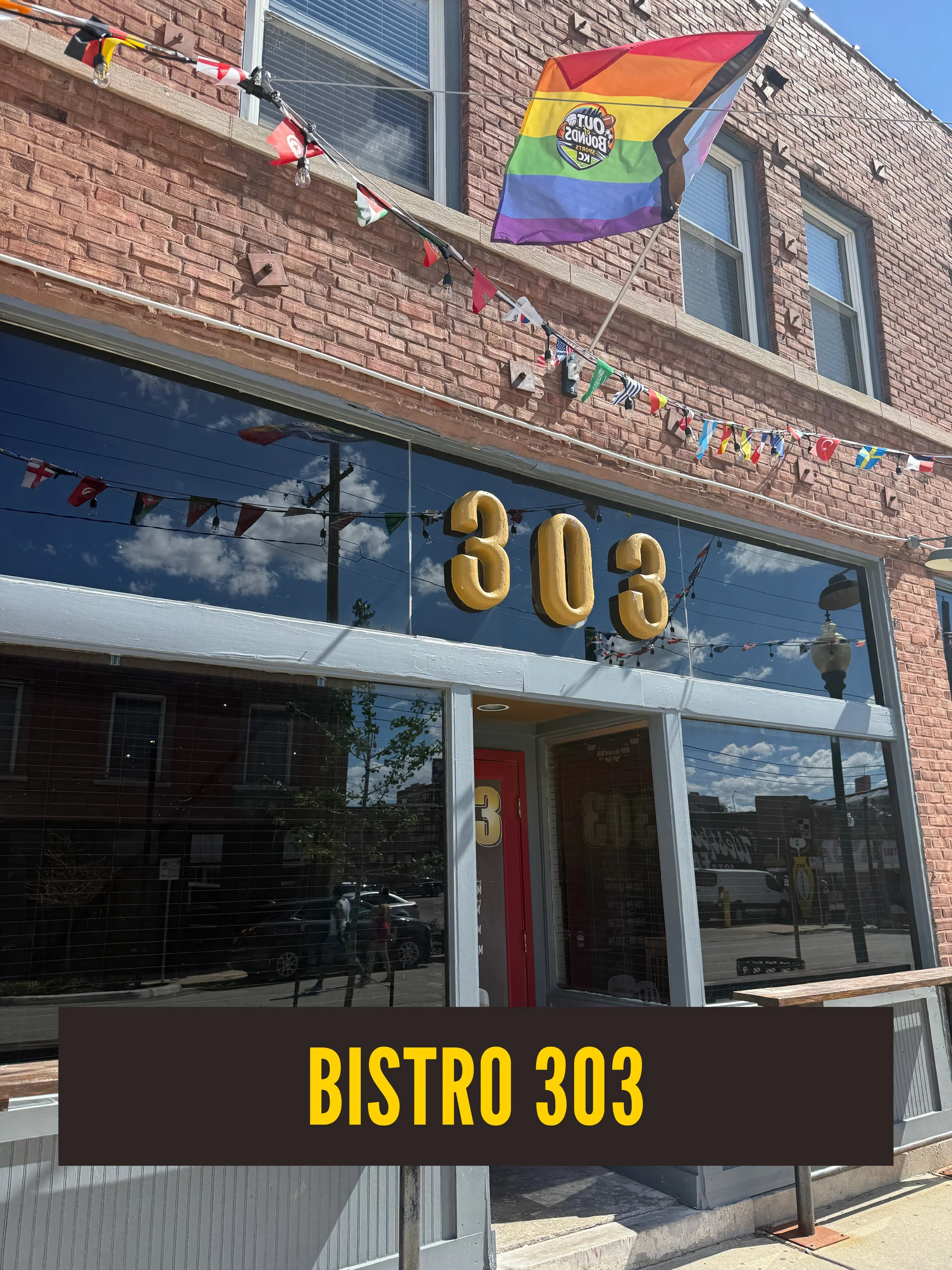 Storefront with large glass windows, the words 'BISTRO 303' in yellow on a black background, and flags including a rainbow pride flag and small international flags hanging on string outside a brick building.  Westport East.