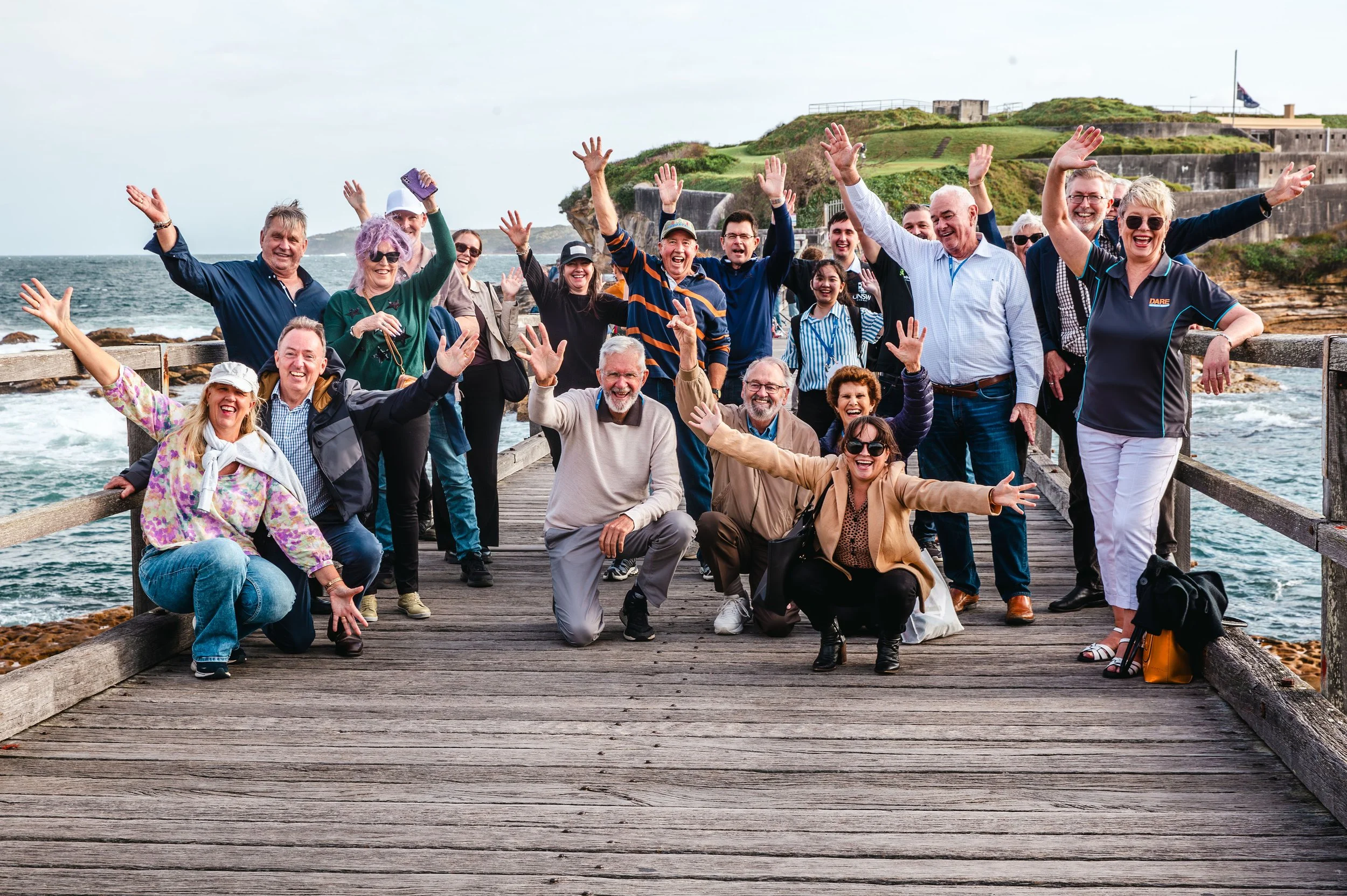 Group of people smiling and waving on a wooden dock by the ocean, with green hills and historical fortification in the background.