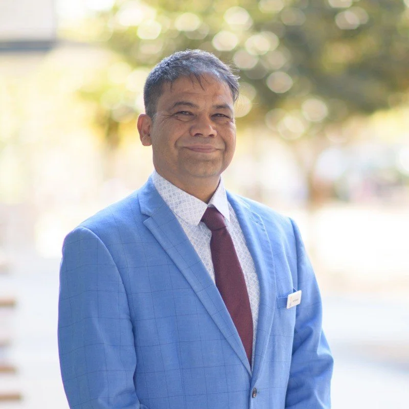 A man in a light blue blazer, white shirt with a subtle pattern, and maroon tie, standing outdoors with blurred trees and sunlight in the background.