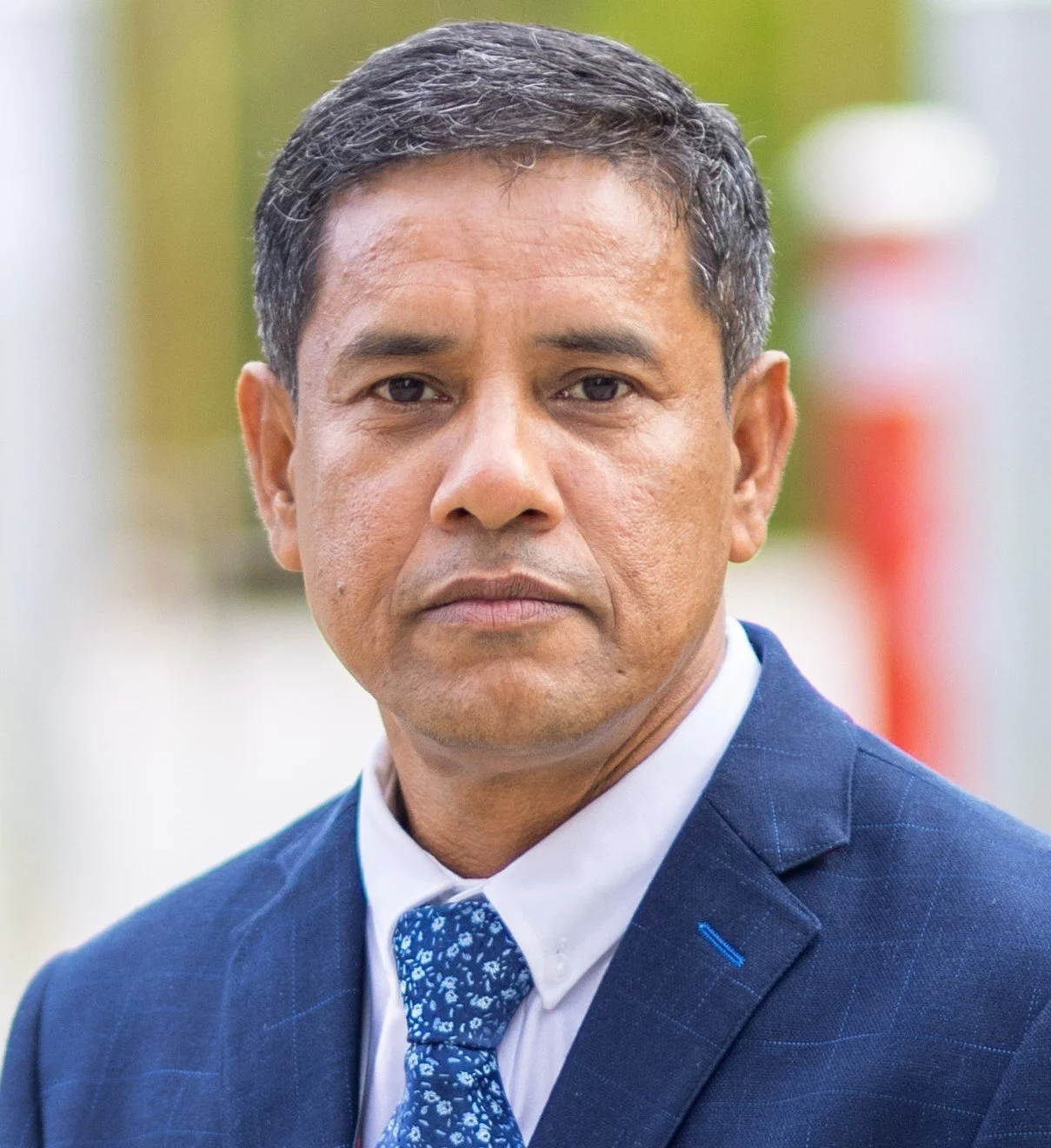 Close-up portrait of a middle-aged man in a suit and tie, outdoors with blurred background.