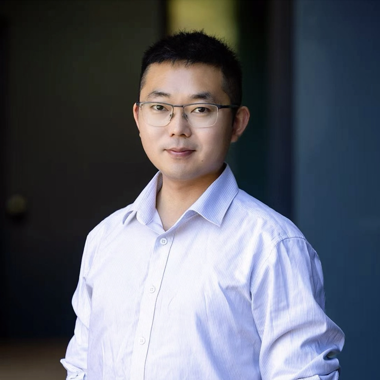 A young man with short black hair, wearing glasses and a light-colored dress shirt, standing outdoors with a blurred background.
