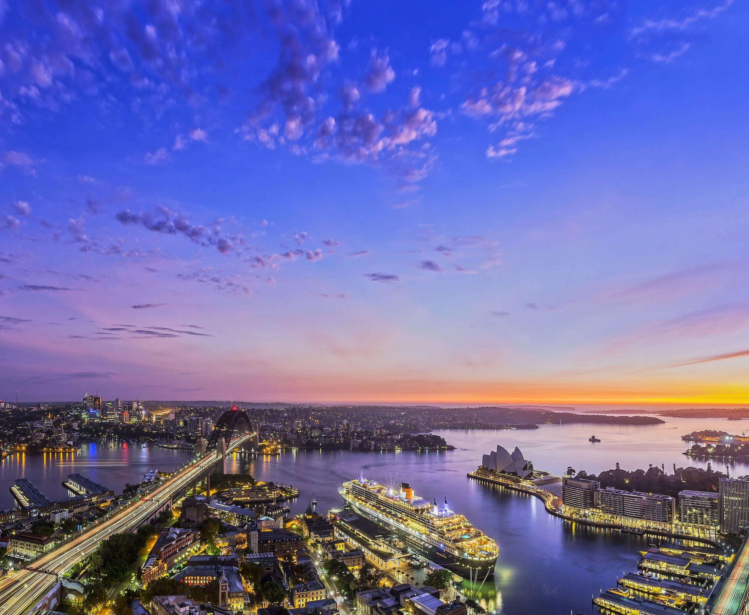 A city skyline at dusk with a river, boats, and illuminated buildings, under a sky with scattered clouds and a colorful sunset.