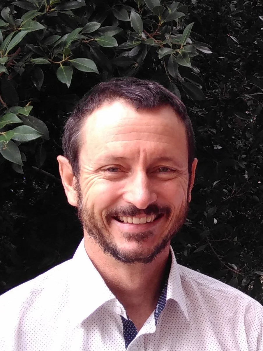 A man with short dark hair and a beard, smiling, wearing a white shirt, standing in front of dense green foliage.