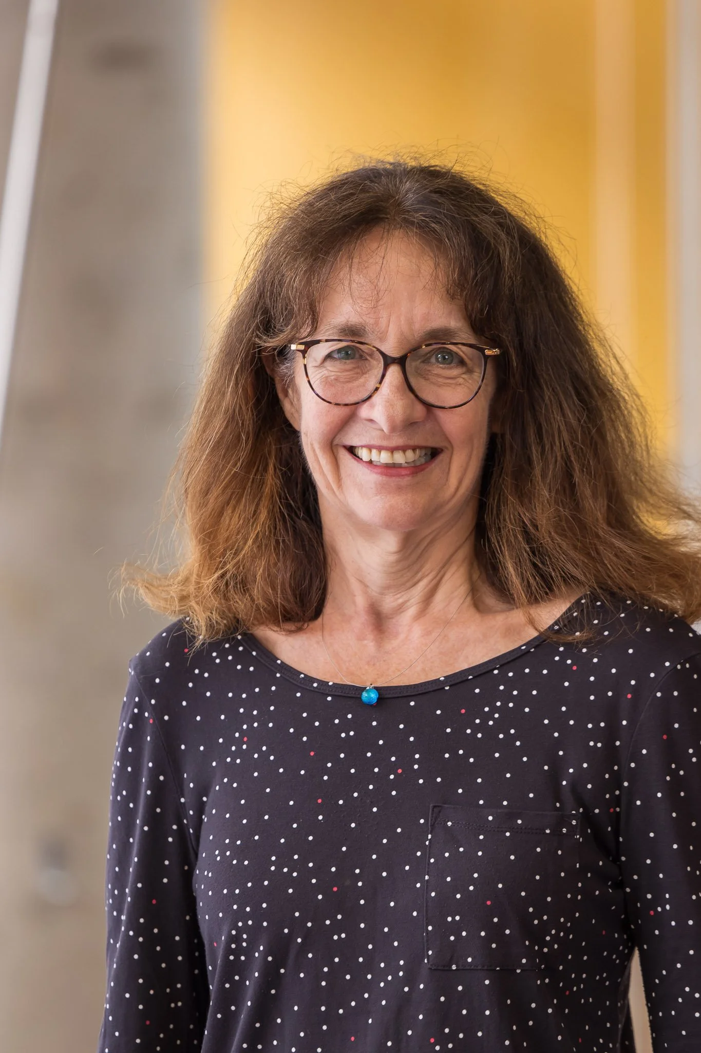 A middle-aged woman with curly brown hair and glasses smiling in a black shirt with white and red polka dots, wearing a blue necklace, standing against a yellow background.