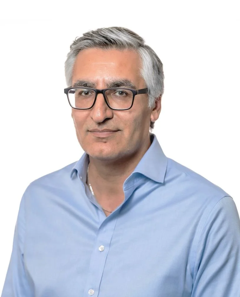 Portrait of a middle-aged man with gray hair wearing black glasses and a light blue collared shirt against a white background.