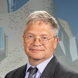 A middle-aged man with gray hair and glasses, wearing a suit and tie, standing in front of a modern building with abstract architectural design.