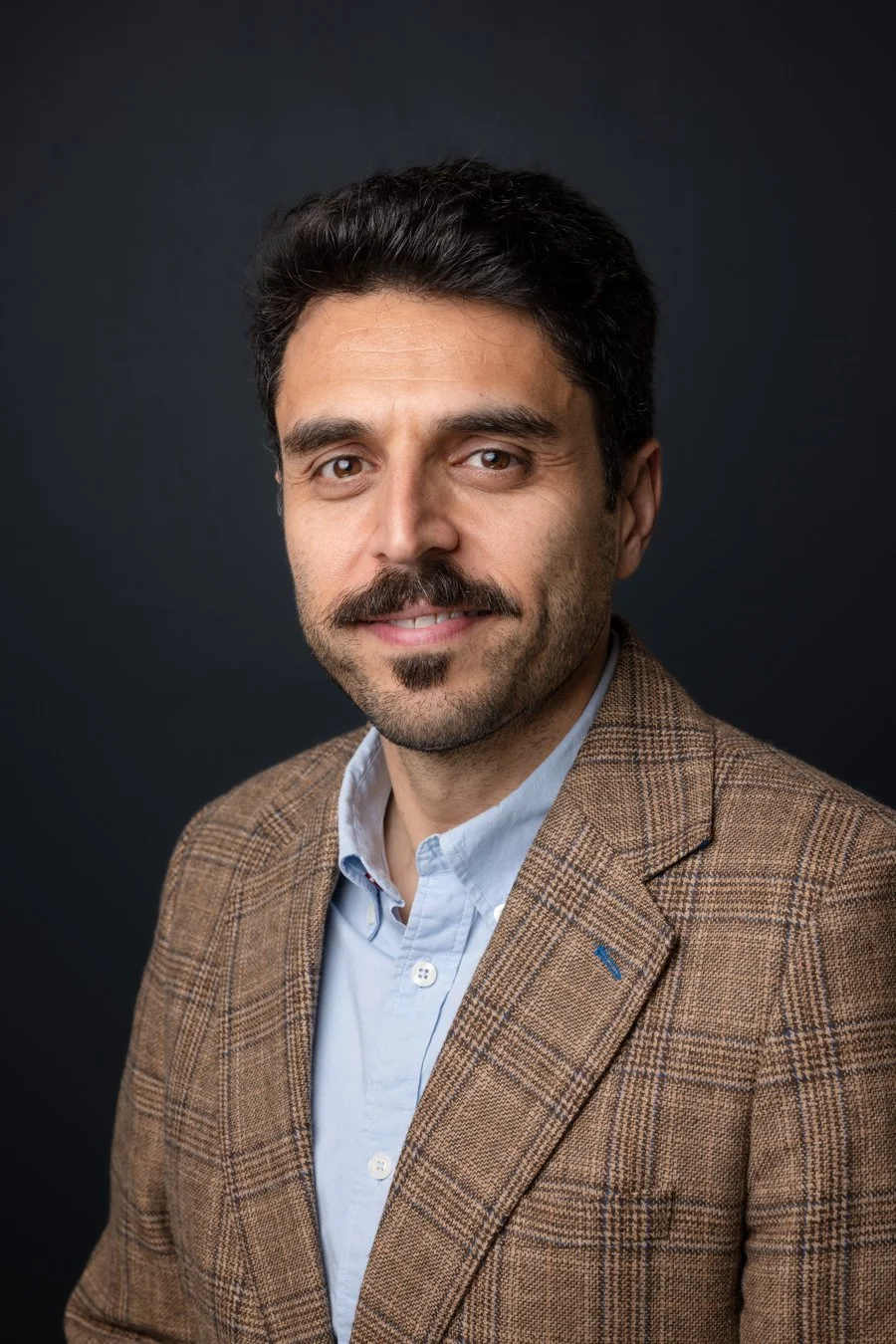 A headshot of a man with dark hair, a beard and mustache, wearing a brown plaid blazer and a light blue collared shirt, against a dark background.