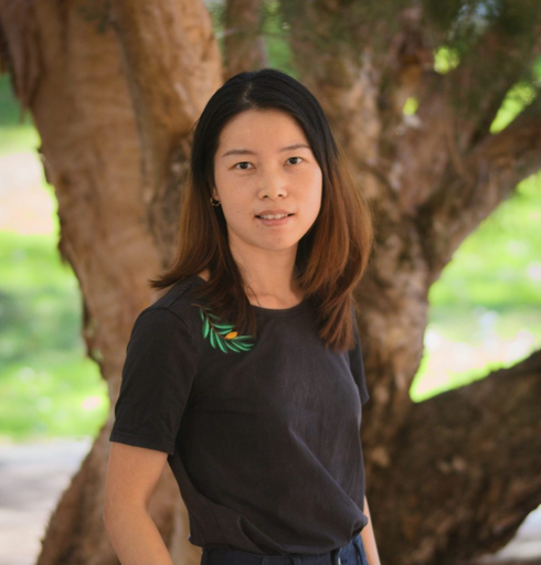 A woman with shoulder-length brown hair standing outdoors in front of a large tree with green foliage.
