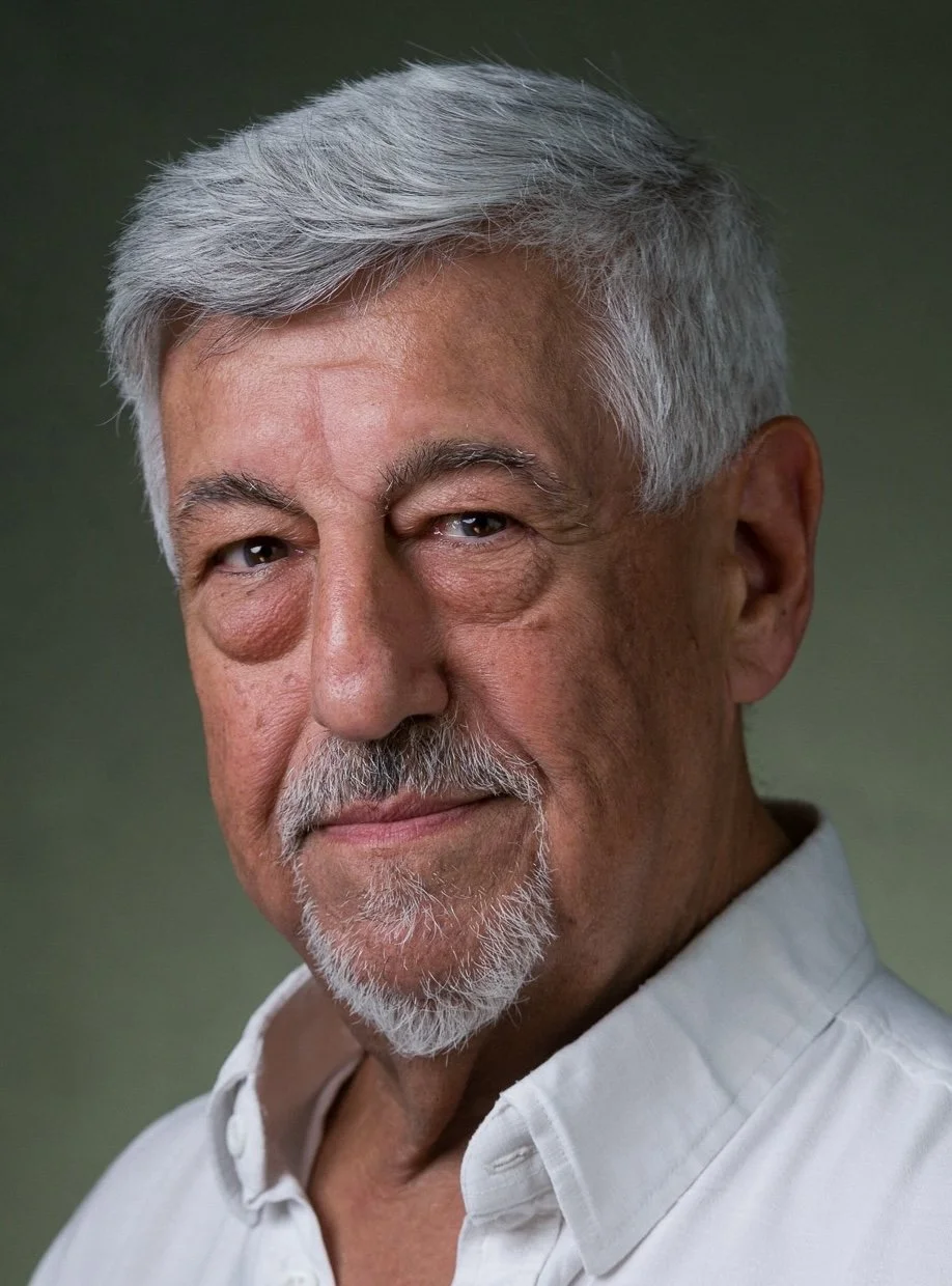 Close-up portrait of an older man with gray hair, beard, and mustache, wearing a white collared shirt, with a neutral expression against a blurred green background.