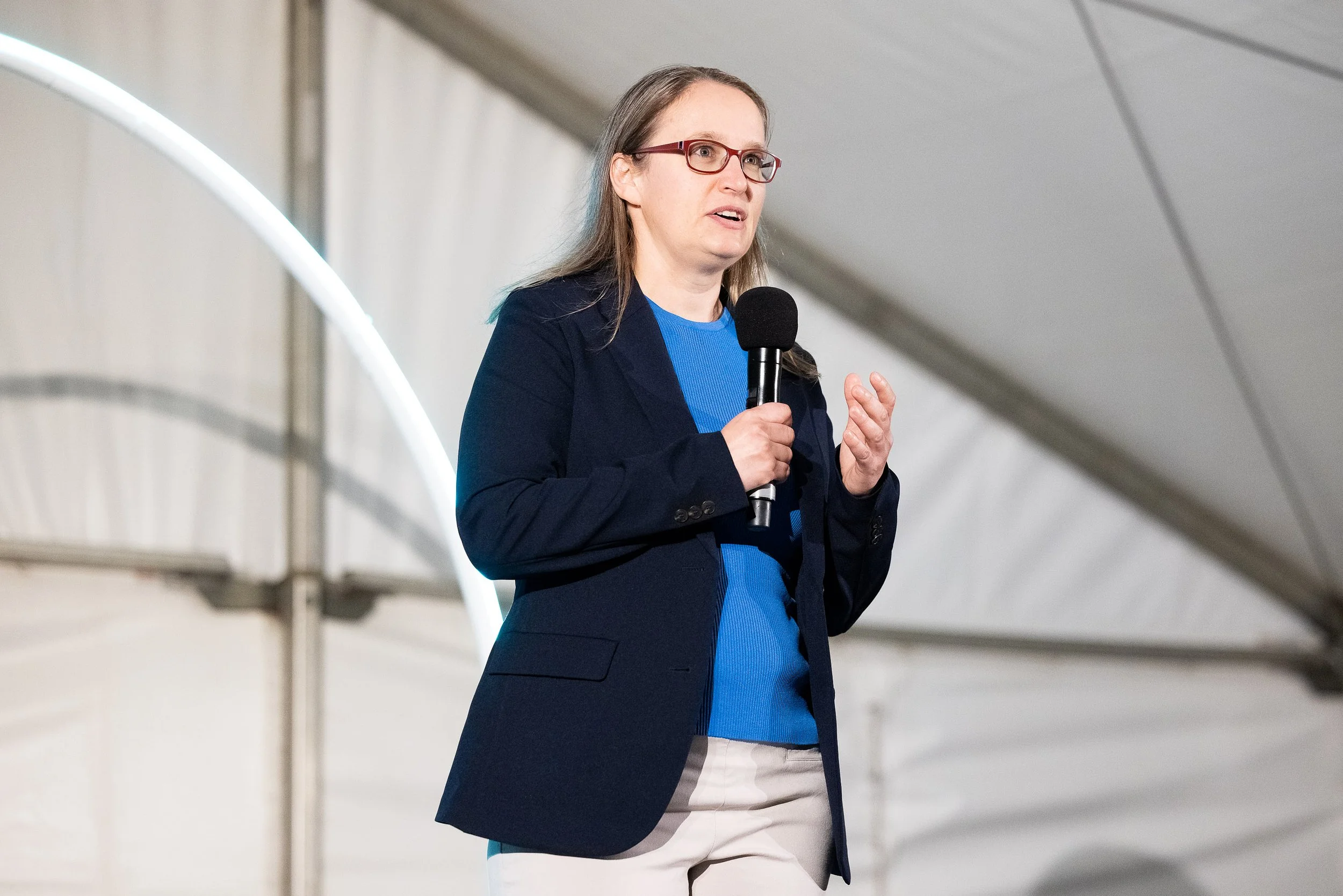 A woman wearing glasses, a navy blazer, a blue shirt, and beige pants is speaking into a microphone during a presentation or speech in an indoor setting.