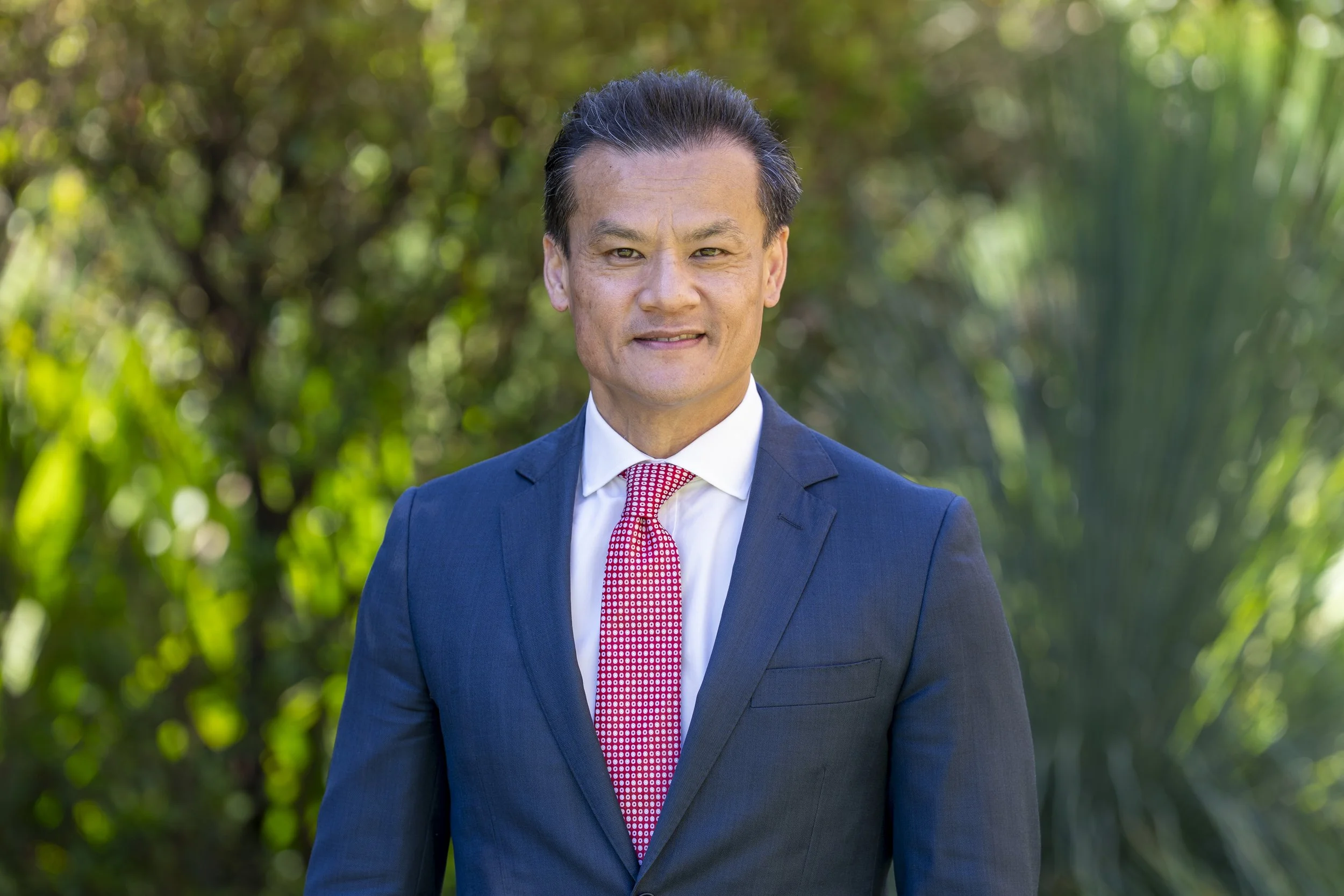 A man in a navy suit with a red tie, standing outdoors with green trees in the background.