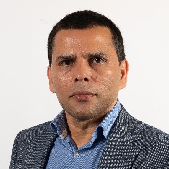 A headshot of a man with dark hair, wearing a blue collared shirt and gray blazer against a plain white background.