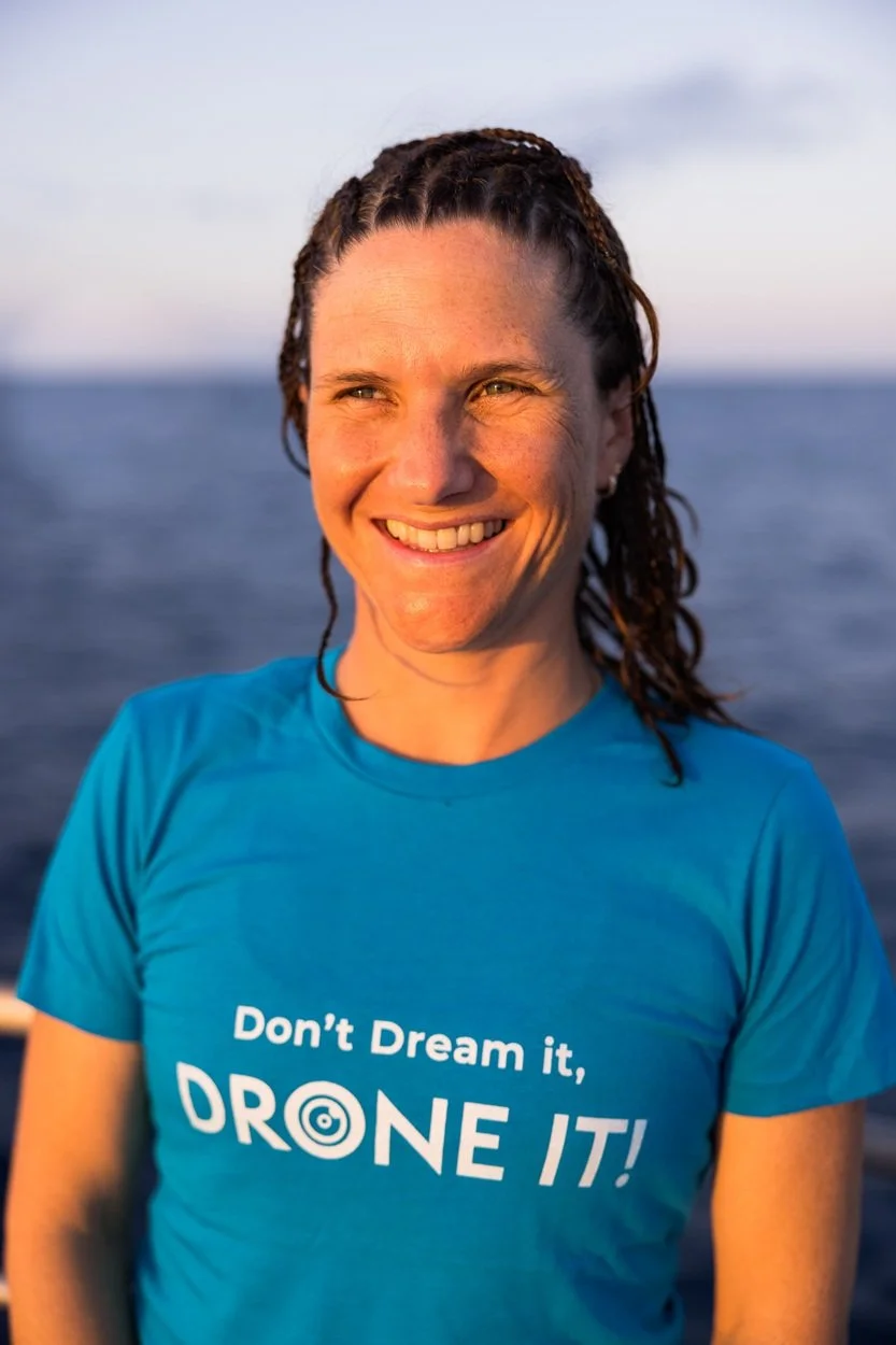A woman with braided hair smiling outdoors near the ocean during sunset, wearing a blue t-shirt that says 'Don’t Dream it, DRONE IT!'