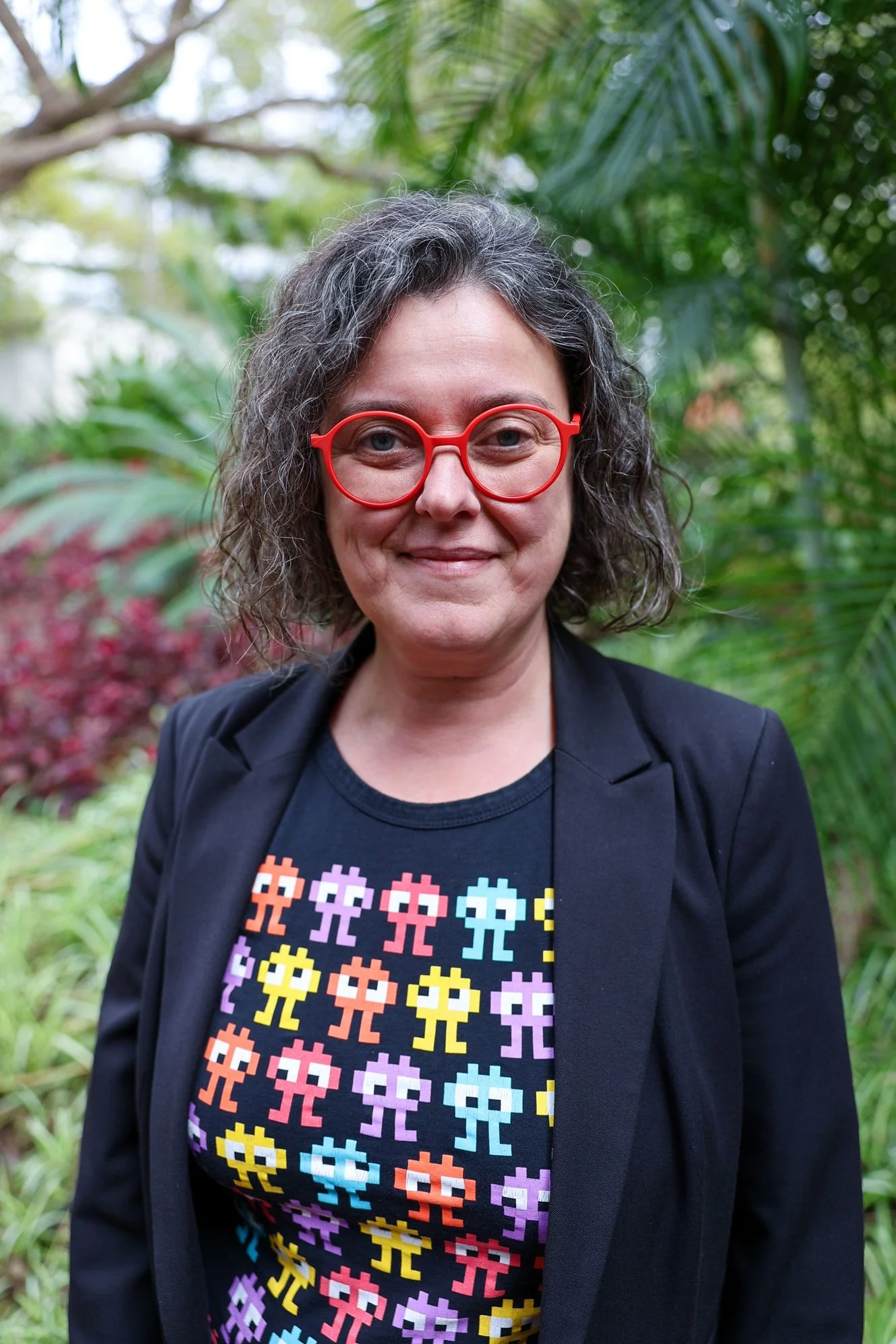 A woman with curly gray hair wearing red glasses and a black jacket stands outdoors in a lush green garden, smiling at the camera.