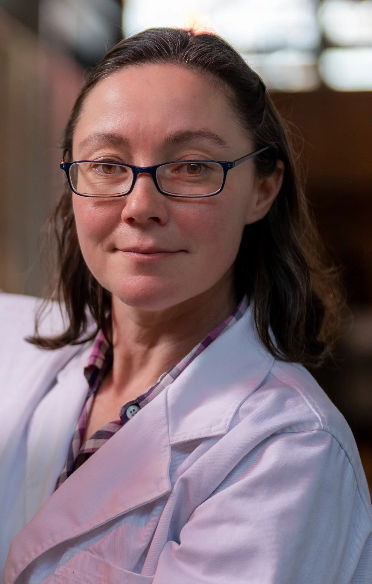 A woman with glasses and brown hair, wearing a white coat, smiling slightly, in a professional setting.