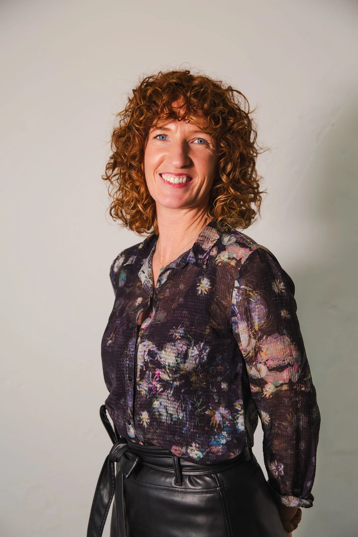 A smiling woman with curly red hair wearing a multicolored floral patterned blouse and black leather pants against a plain wall.